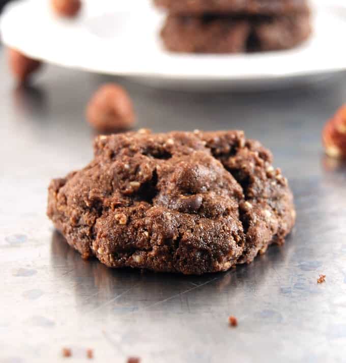 A vegan double chocolate hazelnut cookie on a baking sheet with a few hazelnuts scattered around.