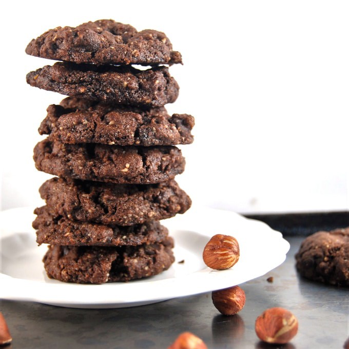 A tall stack of Vegan Chocolate Hazelnut Cookies against a white background with hazelnuts around it