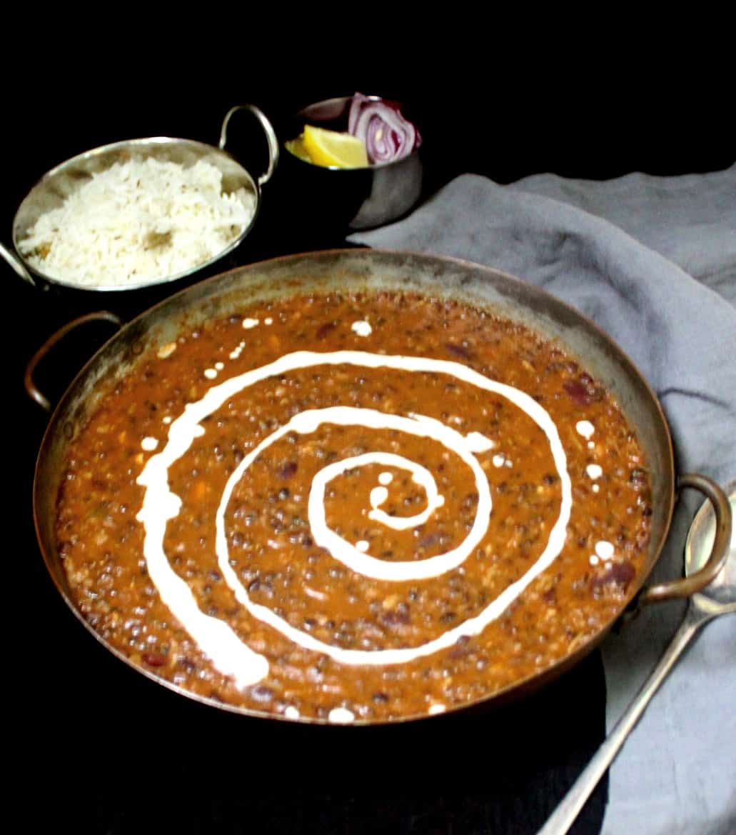 Vegan dal makhani  in a copper serving dish with with cream swirled through it. In the background are cumin rice, slices of onion and lemon, and a gray napkin on a black background.