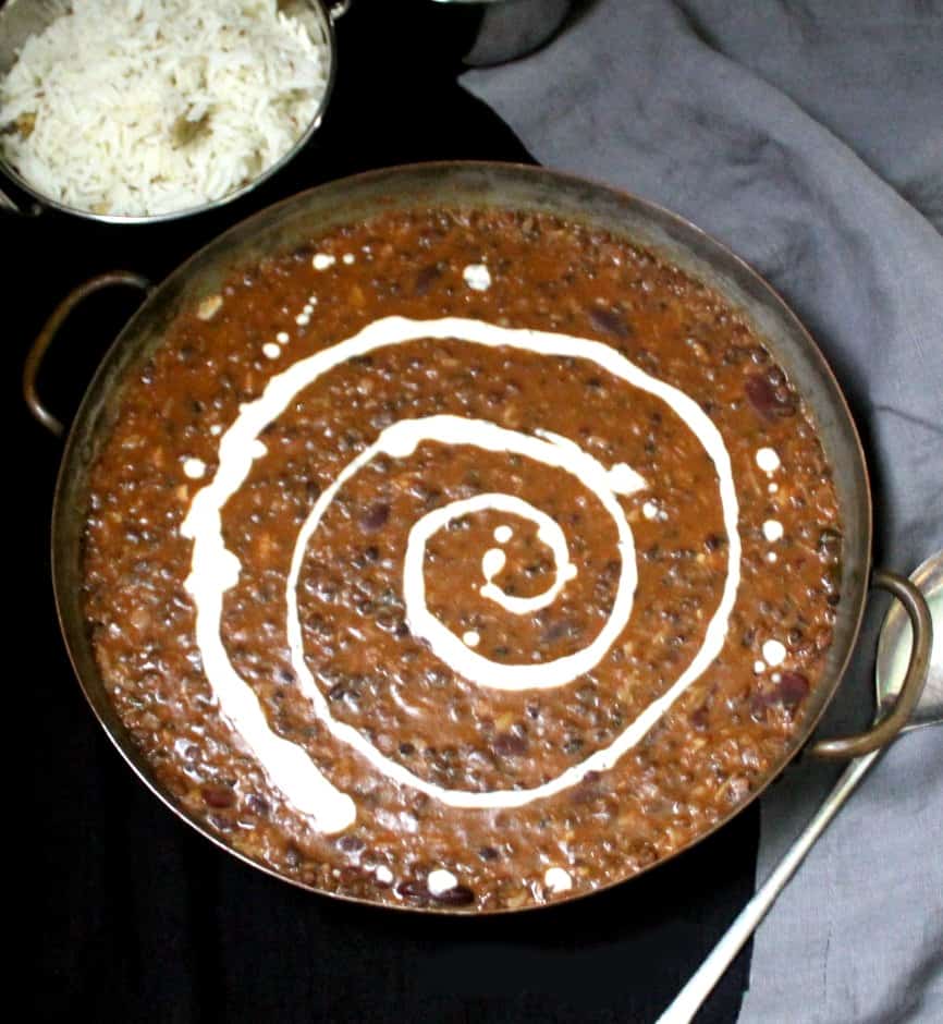 A copper serving dish with creamy and buttery dal makhani with coconot cream swirled through it. In the background are cumin rice, slices of onion and lemon, and a gray napkin on a black background.