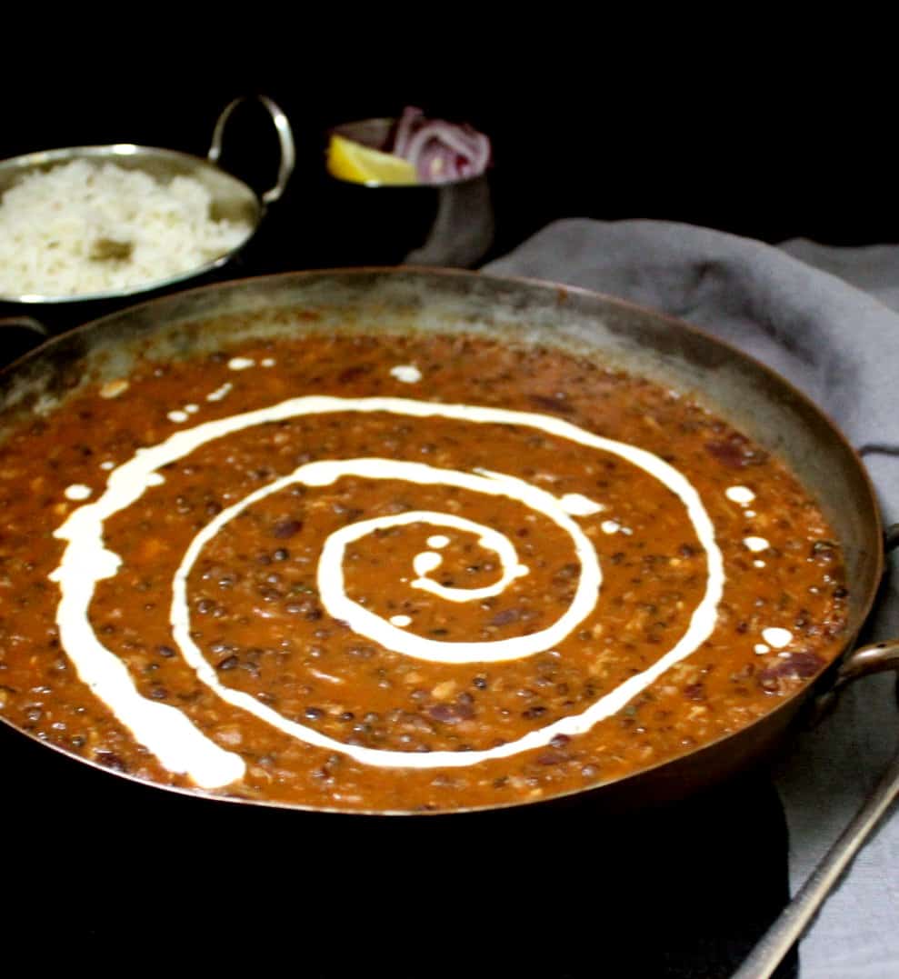 Ccreamy and buttery dal makhani with cashew cream swirled through it. In the background are cumin rice, slices of onion and lemon, and a gray napkin on a black background.