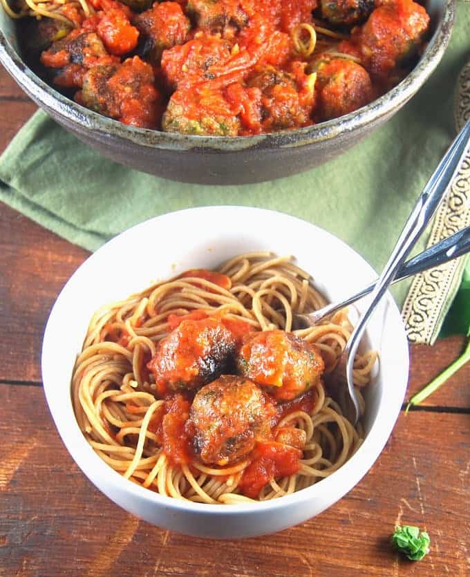 A white bowl with Vegan spaghetti and meatballs, two forks, and a bigger bowl with spaghetti meatballs in background.