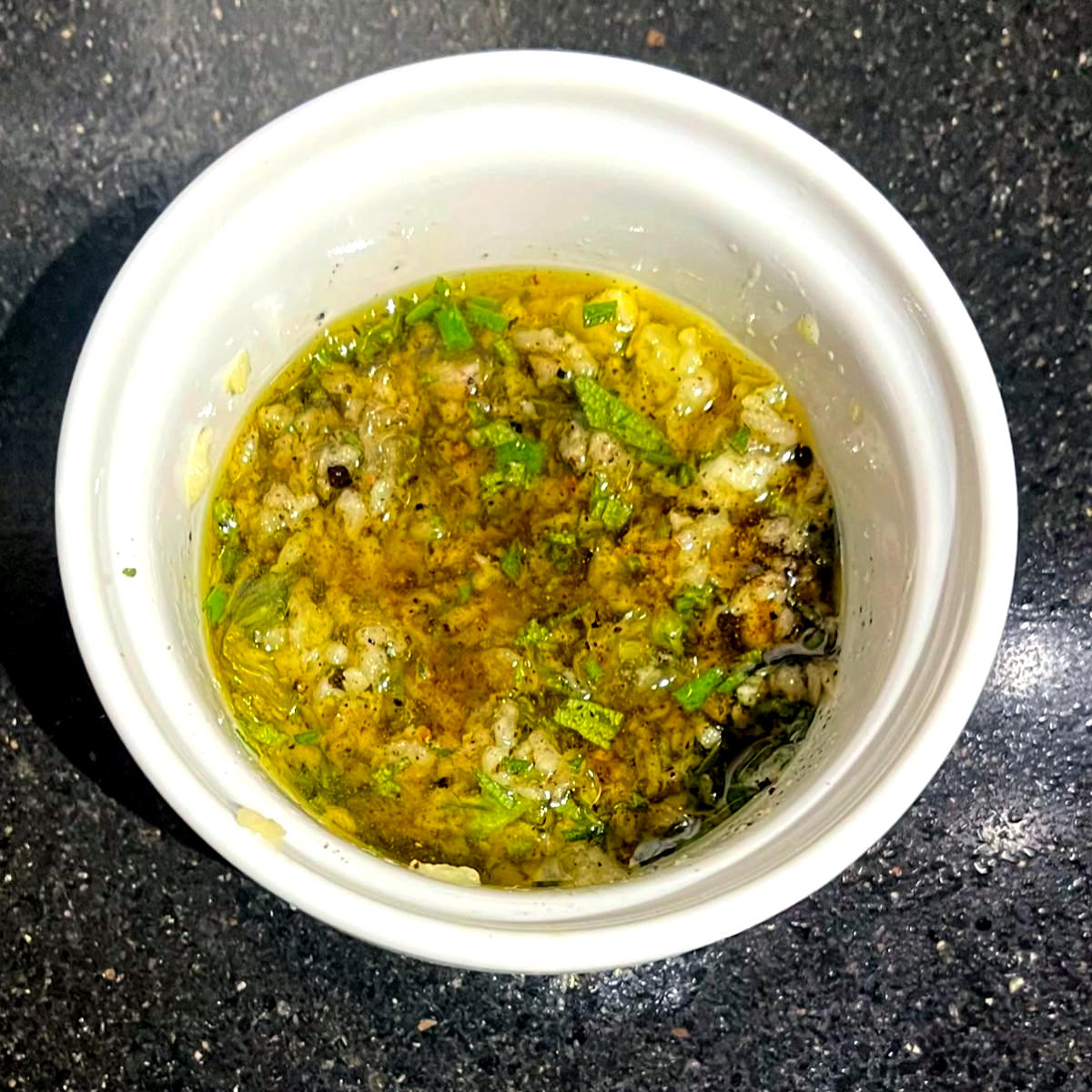 Olive oil with ground black pepper, salt, rosemary and garlic in white ramekin bowl.