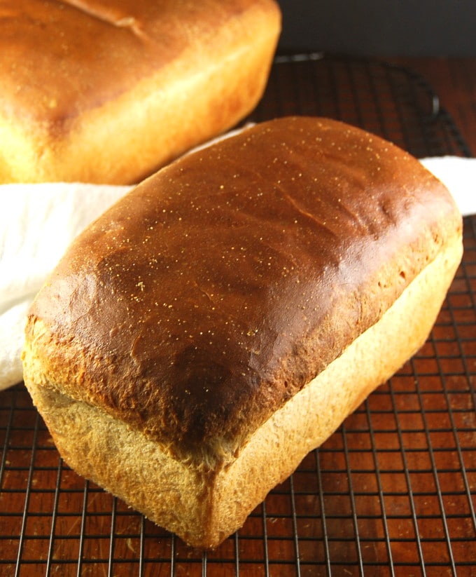 Photo of a loaf of Vegan Sourdough Sandwich Bread on a cooling rack with another loaf in background.