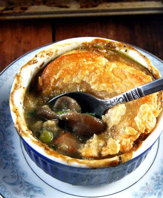 Closeup photo of a mushroom pot pie with the golden puff pastry crust cracked open with a spoon and creamy gravy of mushrooms, peas and carrots peeking through.