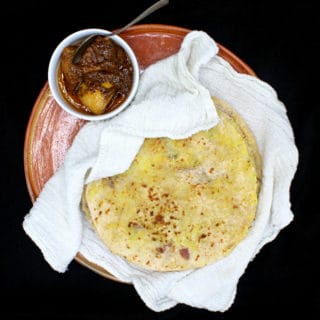 Aloo Parathas stacked on an earthenware dish and nestled inside a white flour sack cloth with a white ceramic bowl of Indian lime pickle on the side with a silver spoon, all on a black background