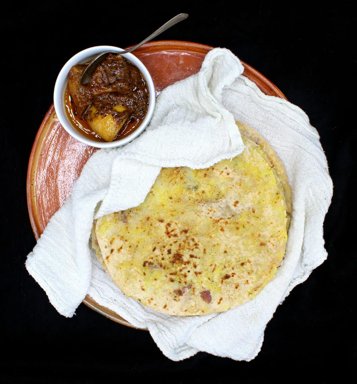 An overhead shot of a stack of freshly baked aloo parathas nestled in a white flour sack towel on an earthenware plate with a small ceramic bowl of Indian lime pickle on the side, all on a black background.