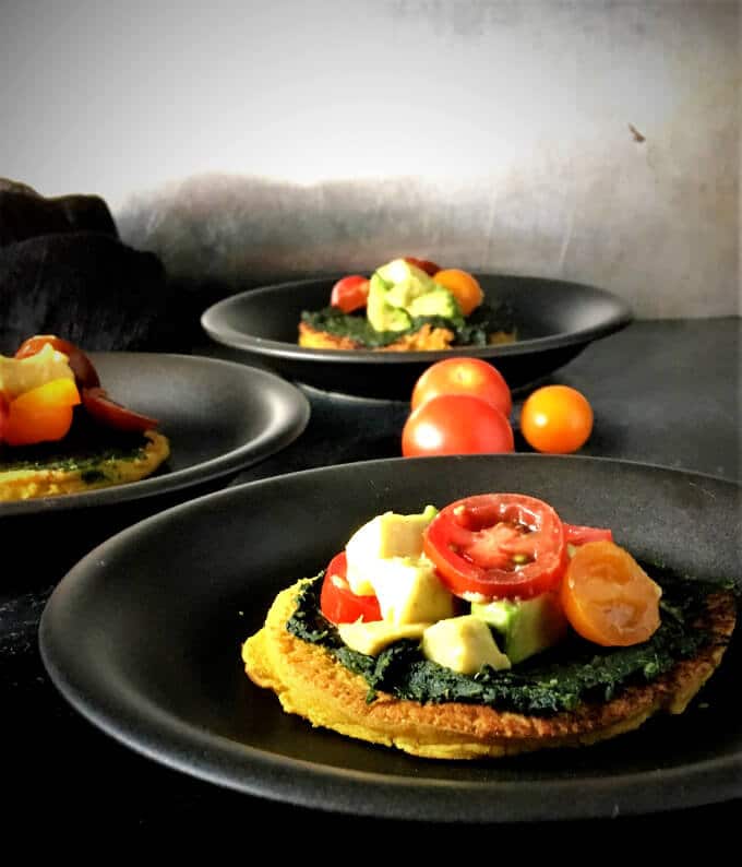 Mini Turmeric Chickpea Crust Pizzas loaded with tomatoes, avocado and spinach on black plates.