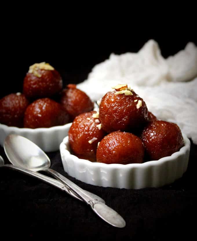 Photo of vegan gulab jamun stacked in white ramekins with a garnish of nuts and spoons next to them.