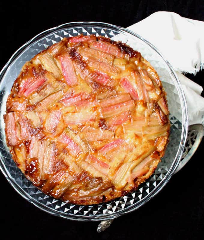 Overhead photo of a vegan rhubarb ginger upside down cake with a soft, moist crumb and a mosaic-like top of glossy pink rhubarb stalks on a glass cake stand.