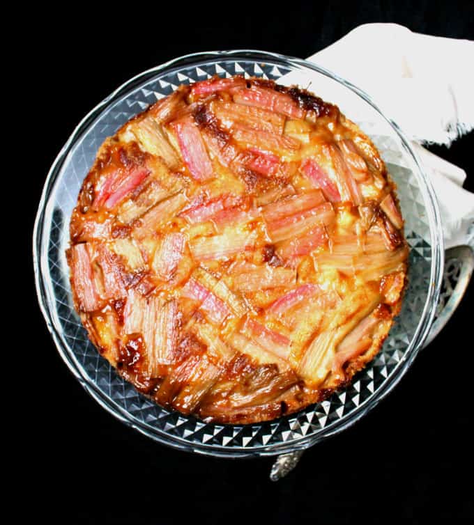 Overhead photo of a rhubarb ginger upside down cake with a soft, moist crumb and a mosaic-like top of glossy pink rhubarb stalks on a glass cake stand.