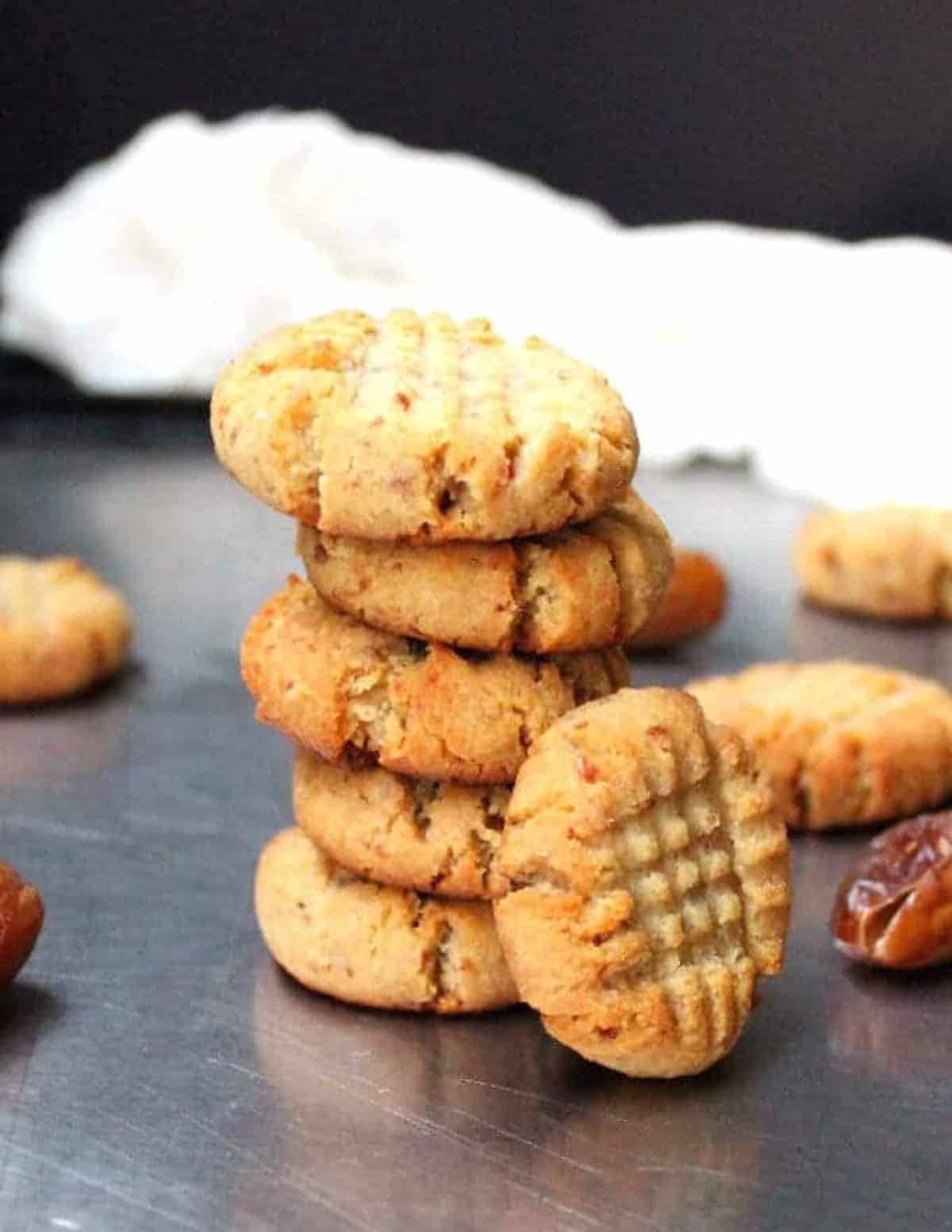 Vegan almond flour cookies stacked on a baking sheet with dates surrounding them.