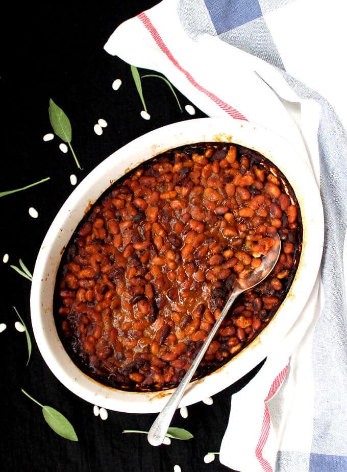Vegan Boston Baked Beans in an oval white baking dish with serving spoon and sage.