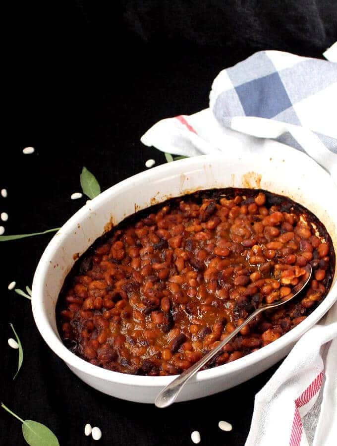 Vegan Boston Baked Beans in an oval white baking dish with serving spoon and sage.