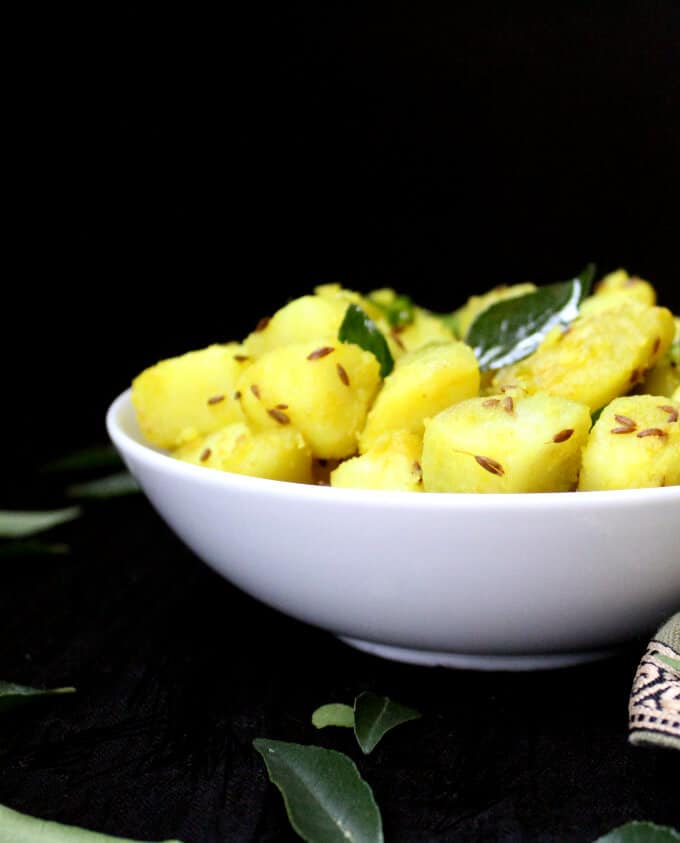 Front partial shot of a white bowl with jeera aloo or cumin potatoes with curry leaves scattered around.