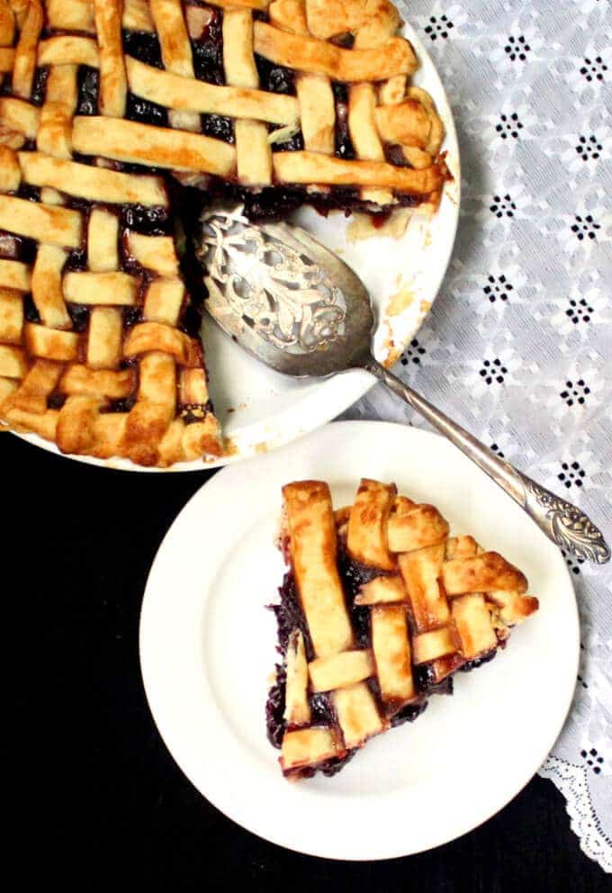 Vegan Cherry Pie slice in white plate and whole pie in background with a silver pie server and lace napkin.