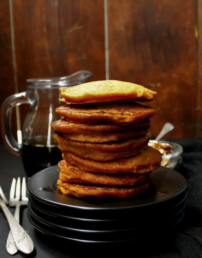 Vegan Carrot Cake Pancakes in a stack of black plates with maple syrup and nuts in the background.