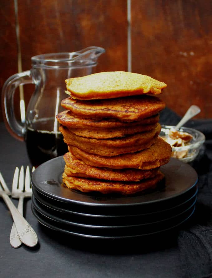 Photo of a stack of Vegan Carrot Cake Pancakes on black plates with maple syrup and nuts in the background.