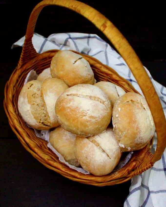 Crusty Sourdough Dinner Rolls, no knead, no added yeast, in a wicker basket with a white and blue kitchen towel on a black background.