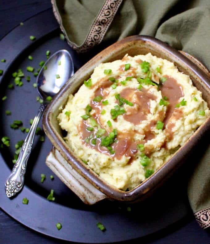Overhead shot of vegan mashed potatoes with gravy in a brown earthenware rectangular bowl with a silver spoon and green napkin.