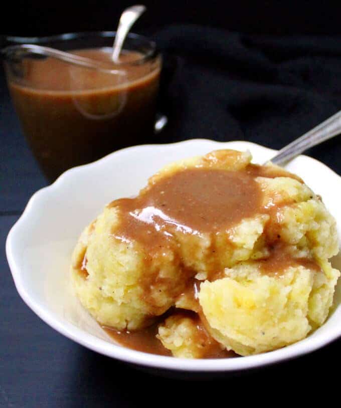 Creamy, rich vegan Mushroom Gravy poured on a bed of creamy vegan mashed potatoes in a white bowl with spoon and in background is a container with more gravy.