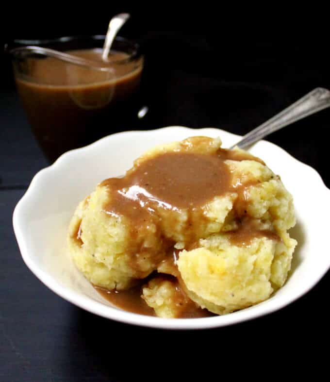 Photo of vegan mushroom gravy served over mashed potatoes in a white bowl with a spoon, with a sauce boat of gravy on the side.