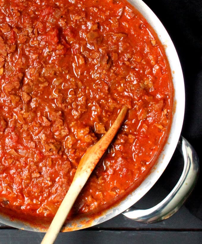 Overhead shot of a wide steel saucepan with meaty vegan marinara sauce and a ladle in it on a gray background.