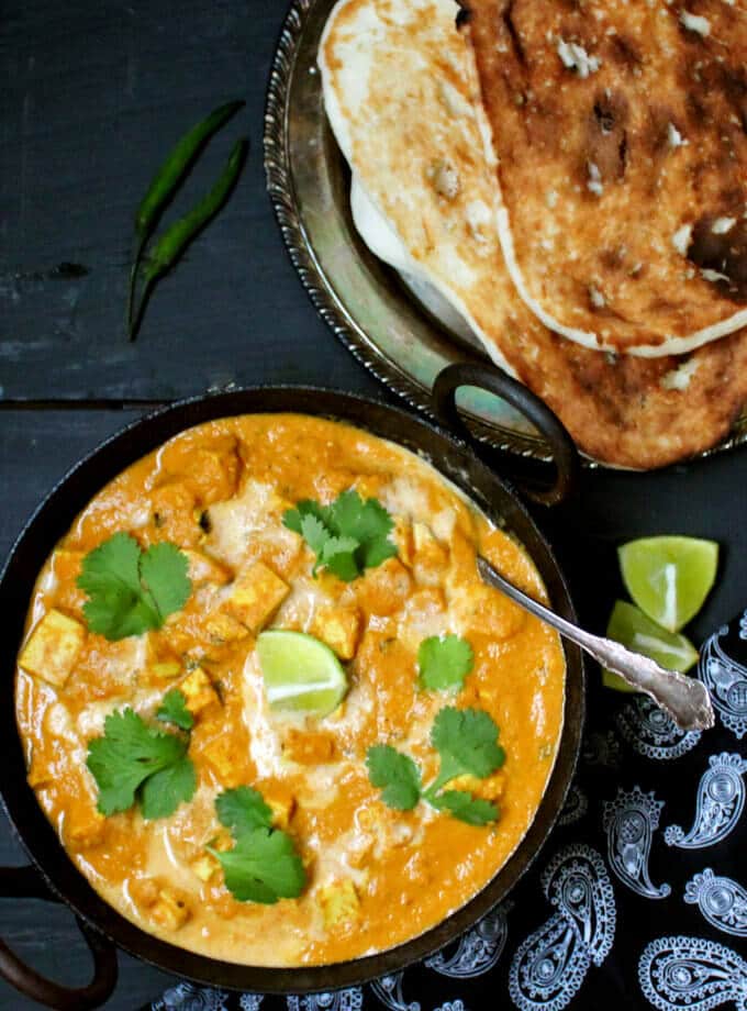 A black kadhai bowl with a fiery orange vegan butter chicken with tofu cubes floating in it, garnished with cashew cream and cilantro. Next to it are vegan naan, wedges of lime, green chili peppers and a black and white paisley print napkin on a black-blue background.