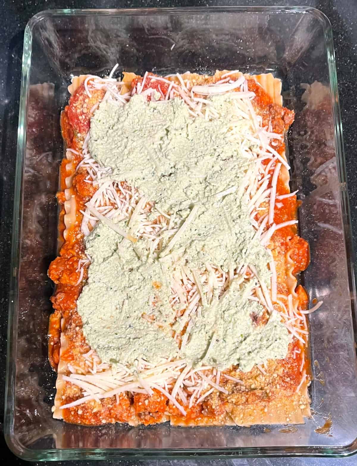 Vegan ricotta layered over vegan parm and tomato sauce and lasagna noodles.