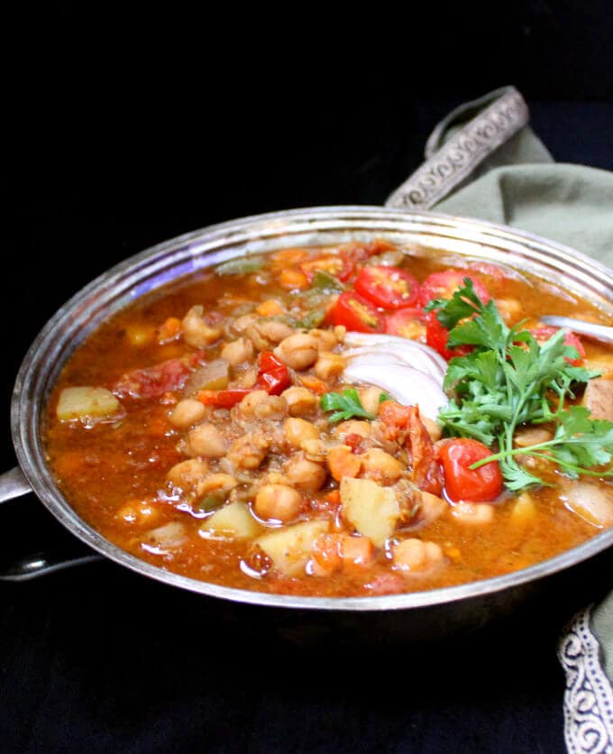An Instant Pot Moroccan Chickpea Stew with no added oil and with chickpeas, tomatoes, carrots and potatoes in a silver server against a black background