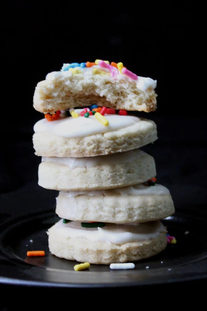 Stack of glutenfree vegan sugar cookies with colorful sprinkles and icing on black plate.