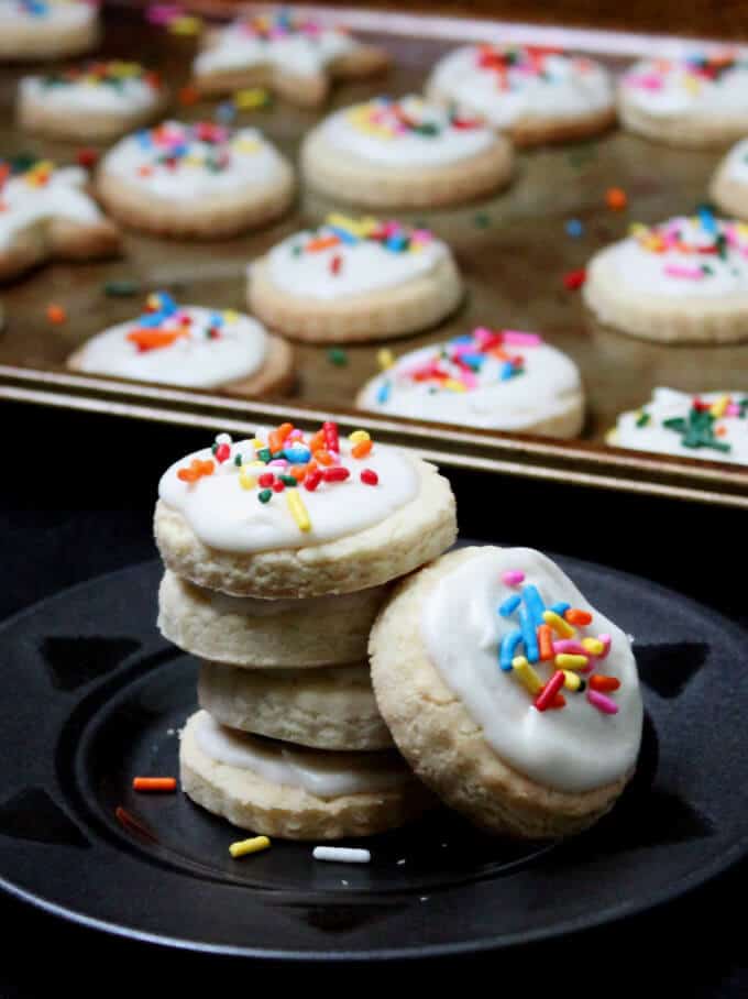 Vegan glutenfree sugar cookies with colorful sprinkles and icing on a black plate with more cookies in baking sheet in background.