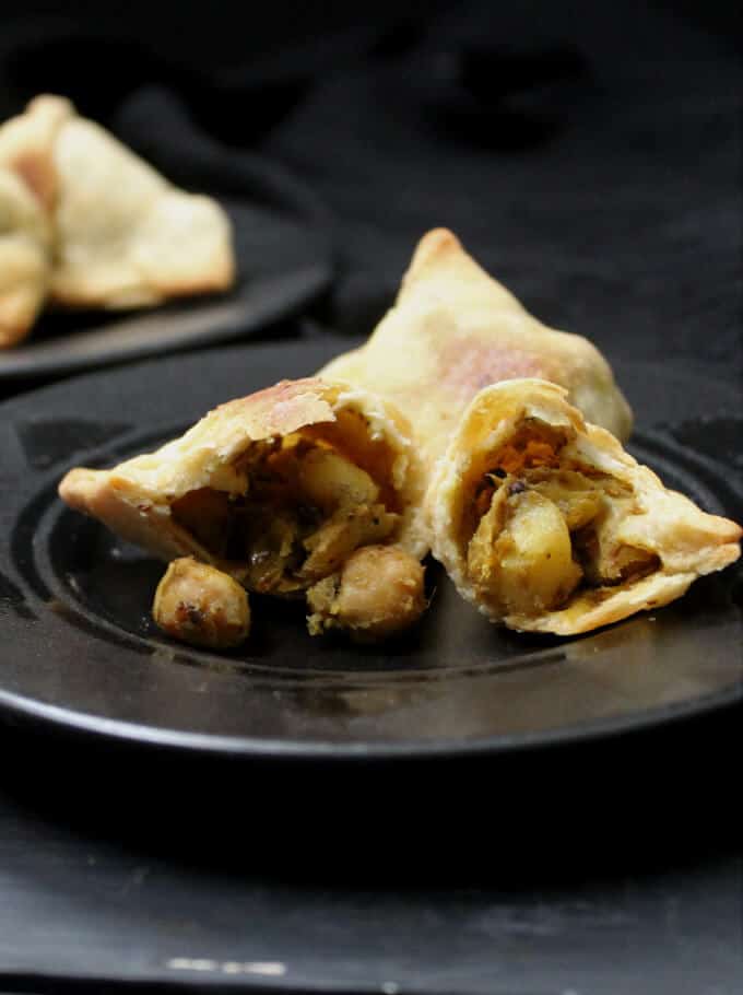 A halved flaky baked samosa with chickpeas and potatoes in the stuffing spilling out on a small black plate