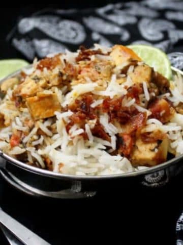 Vegetable biryani with tofu cubes, onions, limes and biryani masala in a steel karahi bowl with a spoon and fork.