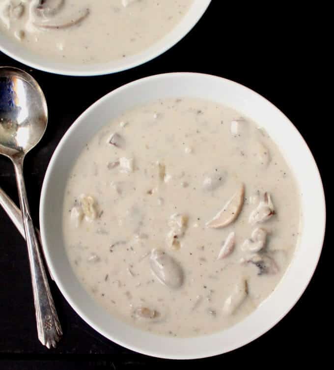 Overhead shot of two white ceramic bowls, one partly visible, with velvety vegan cream of mushroom soup with two silver spoons on a black background