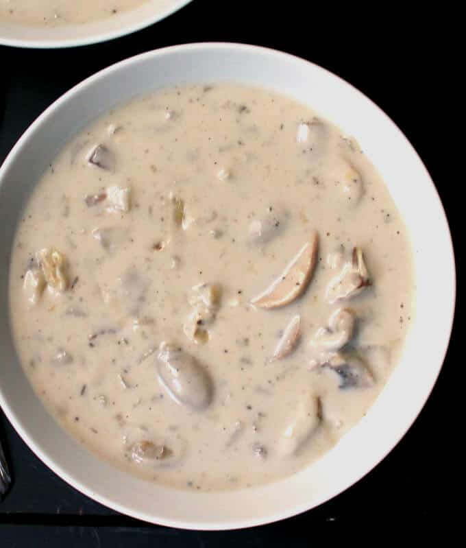 Partial overhead shot of a white bowl with a rich, tasty vegan cream of mushroom soup with bits of mushrooms with a second bowl partially visible at the top on a black background