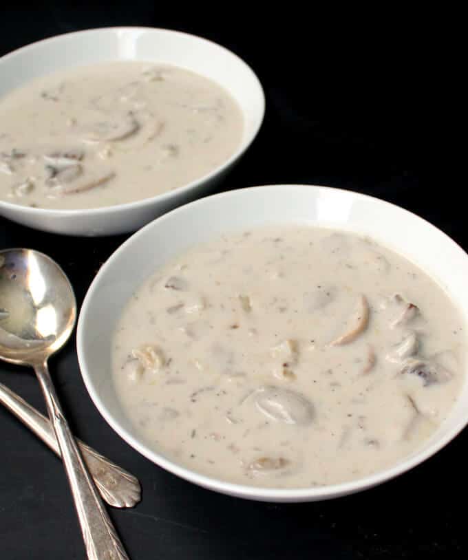 Front shot of two white ceramic bowls of vegan Cream of Mushroom soup with two silver soup spoons on a black background