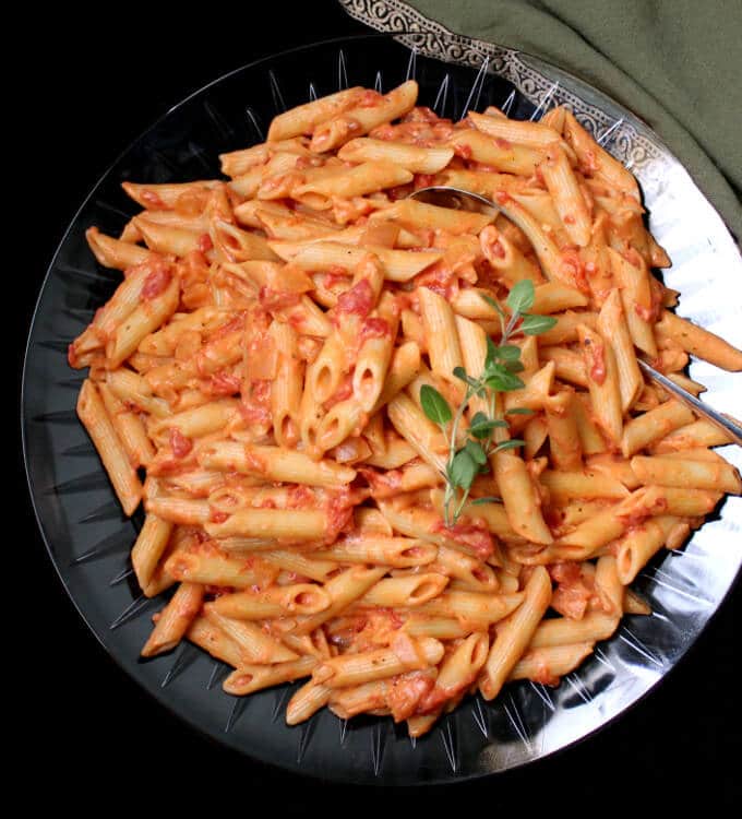 A big crystal platter of vegan penne alla vodka with a spoon, oregano and a green and gold napkin.