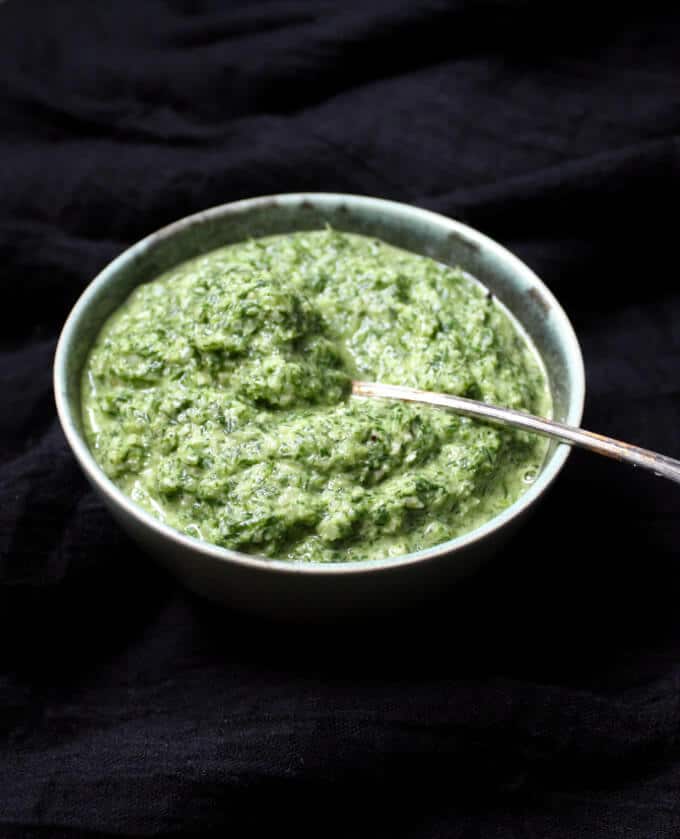 A fresh and green Fennel Fronds Pesto in a blue-green bowl with a silver spoon on a black cheesecloth