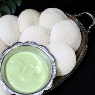 An overhead closeup of fluffy, soft idlis in a silver tray with a bowl of coconut chutney and curry leaves in the background.