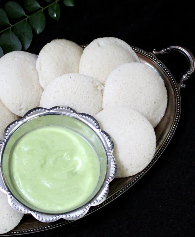 Fluffy, soft idlis in a silver tray with a bowl of coconut chutney and curry leaves in the background.
