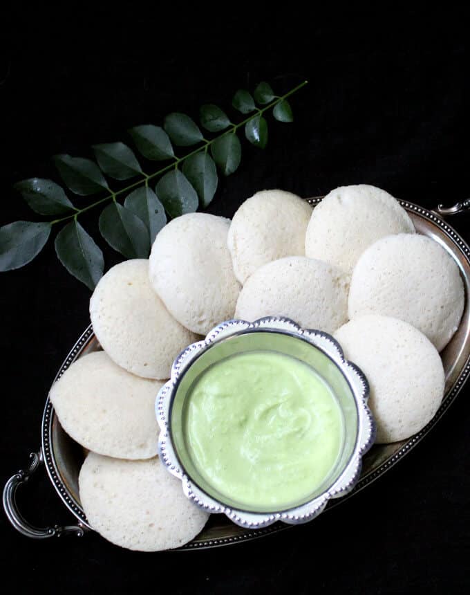 Idli on a silver plate with coconut chutney and curry leaves in background.