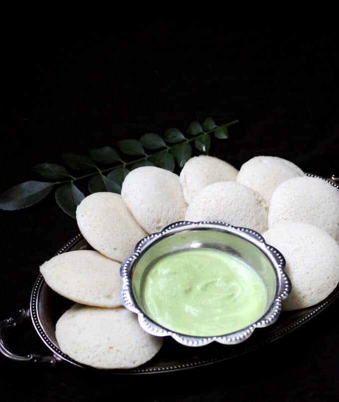 South Indian idli arranged in a silver tray with a bowl of coconut chutney and curry leaves in the background.