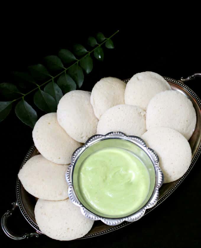 An overhead closeup of fluffy, soft idlis in a silver tray with a bowl of coconut chutney and curry leaves in the background.