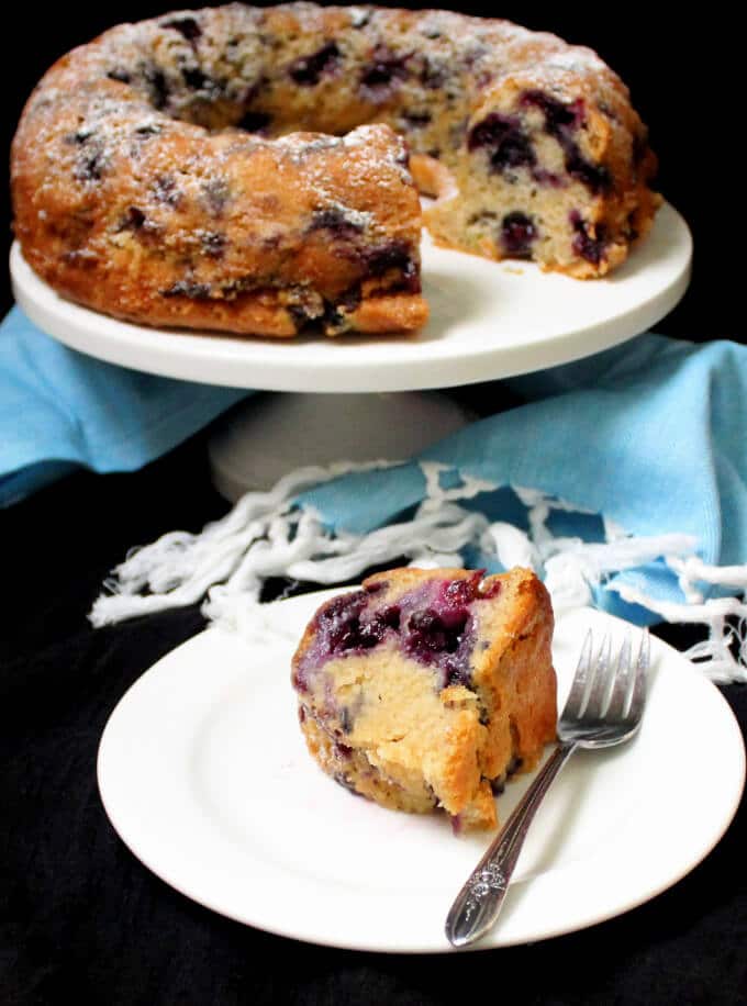 A photo of a blueberry buttermilk bundt cake on a white cake stand witha  slice of the cake on a white plate with a fork and a blue napkin with white tassels.