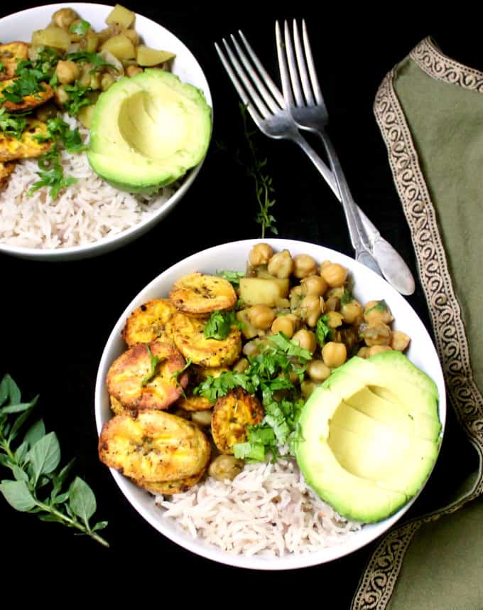 White bowls with plantains, chickpeas, avocado and rice. Forks and a napkin are on the side.