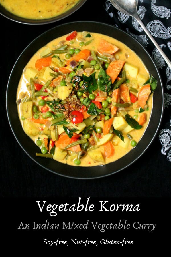 A bowl filled with vegetable korma, made with carrots, potatoes, green beans and tomatoes in a spicy but creamy coconut sauce. Next to the bowl is a paisley napkin and a bowl of dal and spoon.