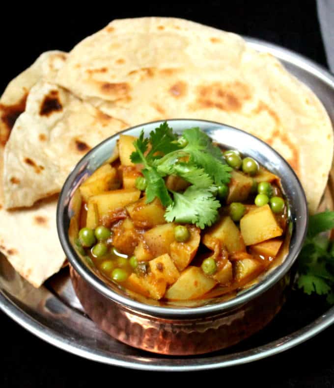 A close up shot of Aloo Matar in a copper and steel handi with rotis in the background on a steel plate