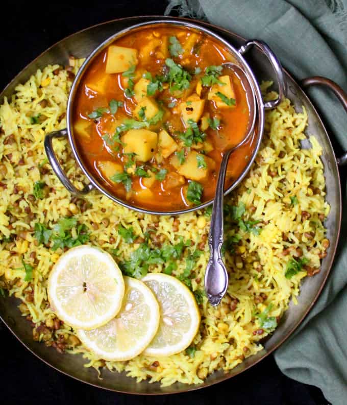 Moong sprouts khichdi in a copper tray with a spicy potato curry against a black background.