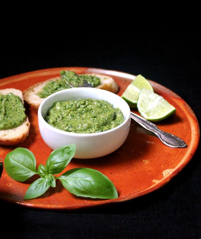 Basil pesto in a white bowl on an earthen plate surrounded by wedges of lime, fresh basil leaves, and slices of bread smeared with pesto for bruschettta.